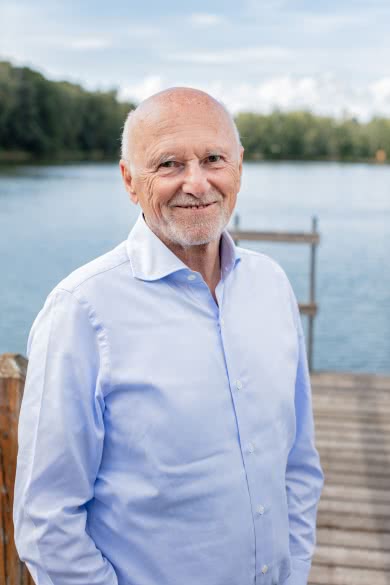 Auf dem Bild sieht man Dirk Rossmann in einem hellblauen Hemd, das vor einem See aufgenommen wurde. Er lächelt leicht in die Kamera.